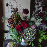 A rustic floral arrangement in a small silver vase on a dark wooden table, featuring deep red roses, green hydrangea, broccoli florets, purple dahlias, and dried botanicals, with a purple kohlrabi bulb in the foreground.