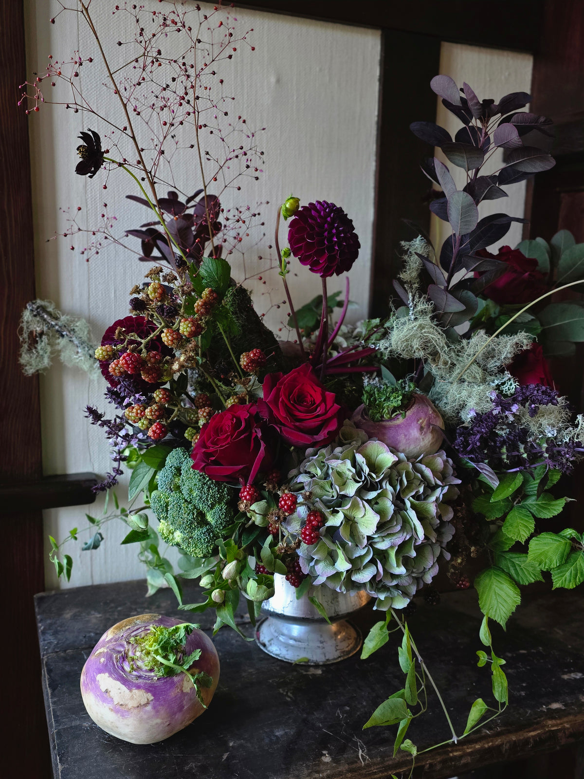 A rustic floral arrangement in a small silver vase on a dark wooden table, featuring deep red roses, green hydrangea, broccoli florets, purple dahlias, and dried botanicals, with a purple kohlrabi bulb in the foreground.