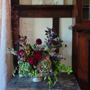 A rustic floral arrangement in a small silver-colored metal vase, featuring deep red roses, burgundy hydrangea, green foliage, and purple accents, set on a dark wooden table in front of a wooden chair.