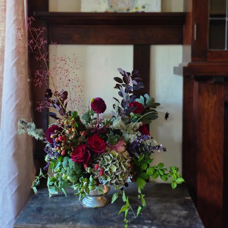 A rustic floral arrangement in a small silver-colored metal vase, featuring deep red roses, burgundy hydrangea, green foliage, and purple accents, set on a dark wooden table in front of a wooden chair.