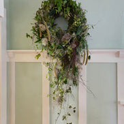 A lush, multi-textured green botanical wreath hangs on a pale sage green wall above white wainscoting, with trailing vines and dried elements cascading down.