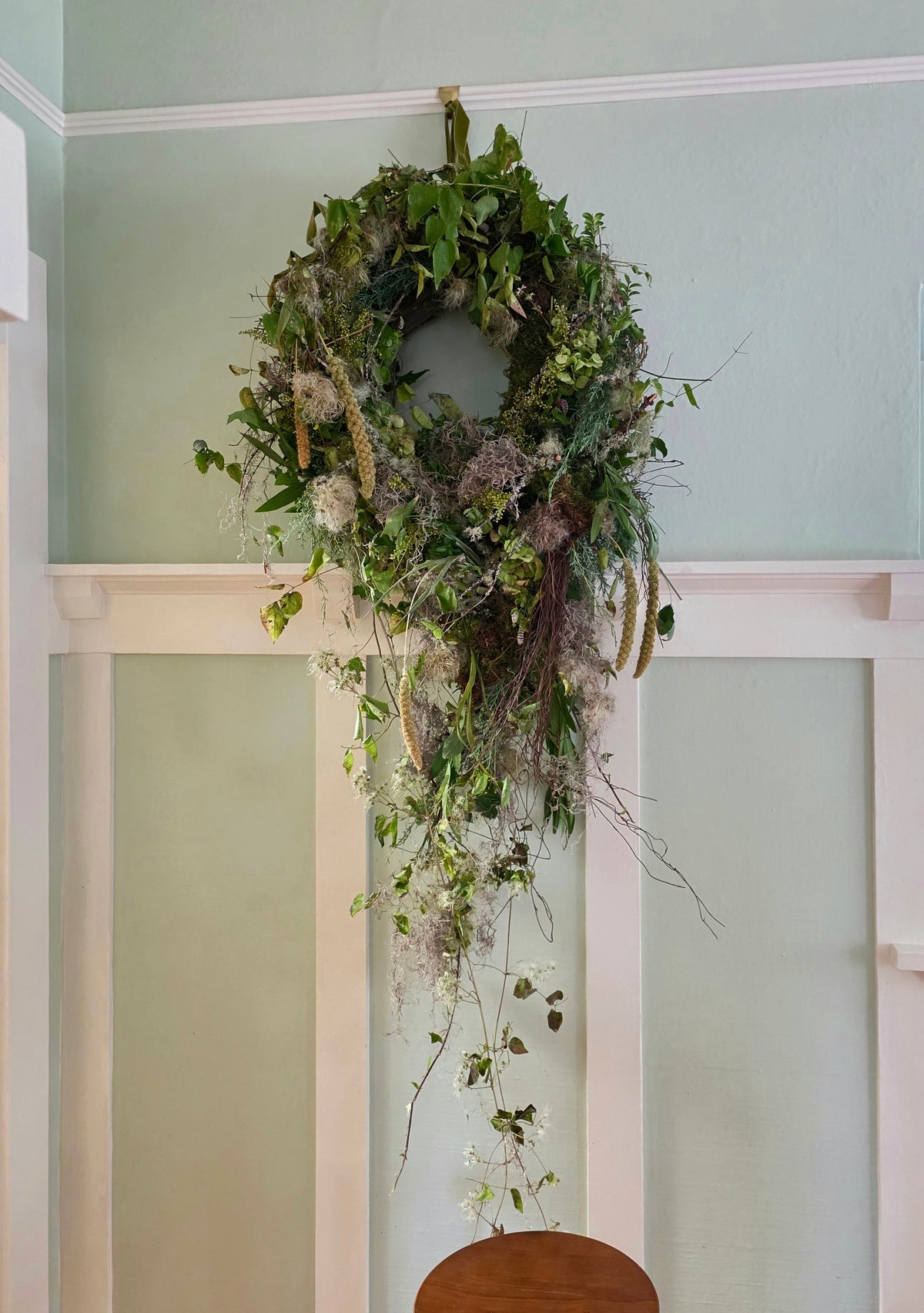 A lush, multi-textured green botanical wreath hangs on a pale sage green wall above white wainscoting, with trailing vines and dried elements cascading down.
