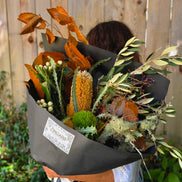 A person holding a large dried flower bouquet wrapped in black paper, with a visible 'DREAMS' label, set against a wooden fence and green garden background.