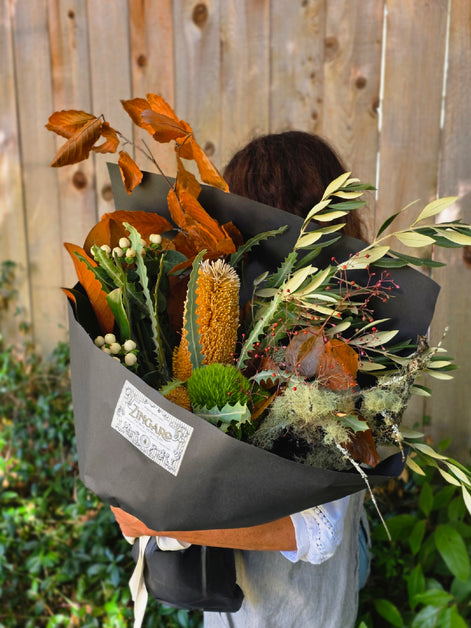 A person holding a large dried flower bouquet wrapped in black paper, with a visible 'DREAMS' label, set against a wooden fence and green garden background.