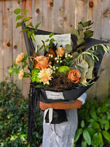 A person holding a large, beautifully arranged floral bouquet wrapped in black paper. The bouquet features orange roses, peach chrysanthemums, green foliage, and small white filler flowers.