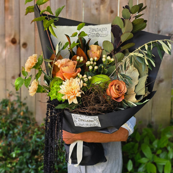 A person holding a large, beautifully arranged floral bouquet wrapped in black paper. The bouquet features orange roses, peach chrysanthemums, green foliage, and small white filler flowers.