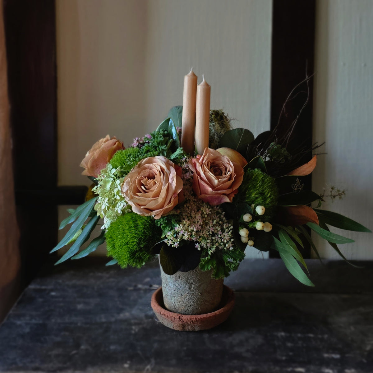 A rustic floral centerpiece in a small terracotta pot, featuring peach roses, green moss, eucalyptus leaves, and two tall beige taper candles, set on a dark wooden surface.