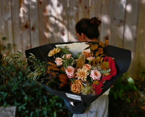 A person viewed from behind holding a large, professionally wrapped flower bouquet with autumnal tones of orange, pink, red, and green foliage, set against a wooden fence background.