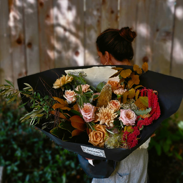 A person viewed from behind holding a large, professionally wrapped flower bouquet with autumnal tones of orange, pink, red, and green foliage, set against a wooden fence background.