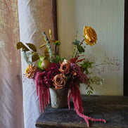 A rustic floral arrangement in a distressed ceramic pot, featuring yellow and peach roses, burgundy berries, and green foliage, displayed on a dark wooden table next to a sheer curtain.