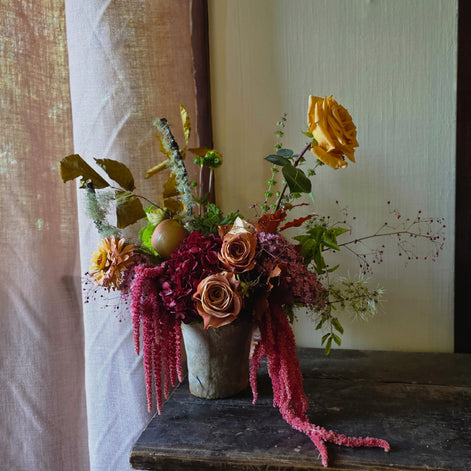 A rustic floral arrangement in a distressed ceramic pot, featuring yellow and peach roses, burgundy berries, and green foliage, displayed on a dark wooden table next to a sheer curtain.