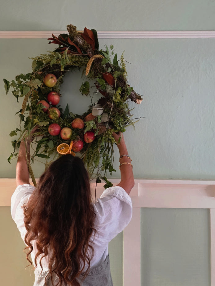 A woman with long wavy brown hair, seen from behind, hangs a large decorative wreath on a pale green wall. The wreath is made of green foliage, brown branches, and red apples, with a dried orange slice accent.
