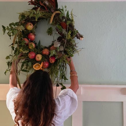 A woman with long wavy brown hair, seen from behind, hangs a large decorative wreath on a pale green wall. The wreath is made of green foliage, brown branches, and red apples, with a dried orange slice accent.