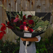 A large, beautifully arranged dried and fresh flower bouquet wrapped in black paper, held by a person wearing a gray apron in front of a wooden fence.