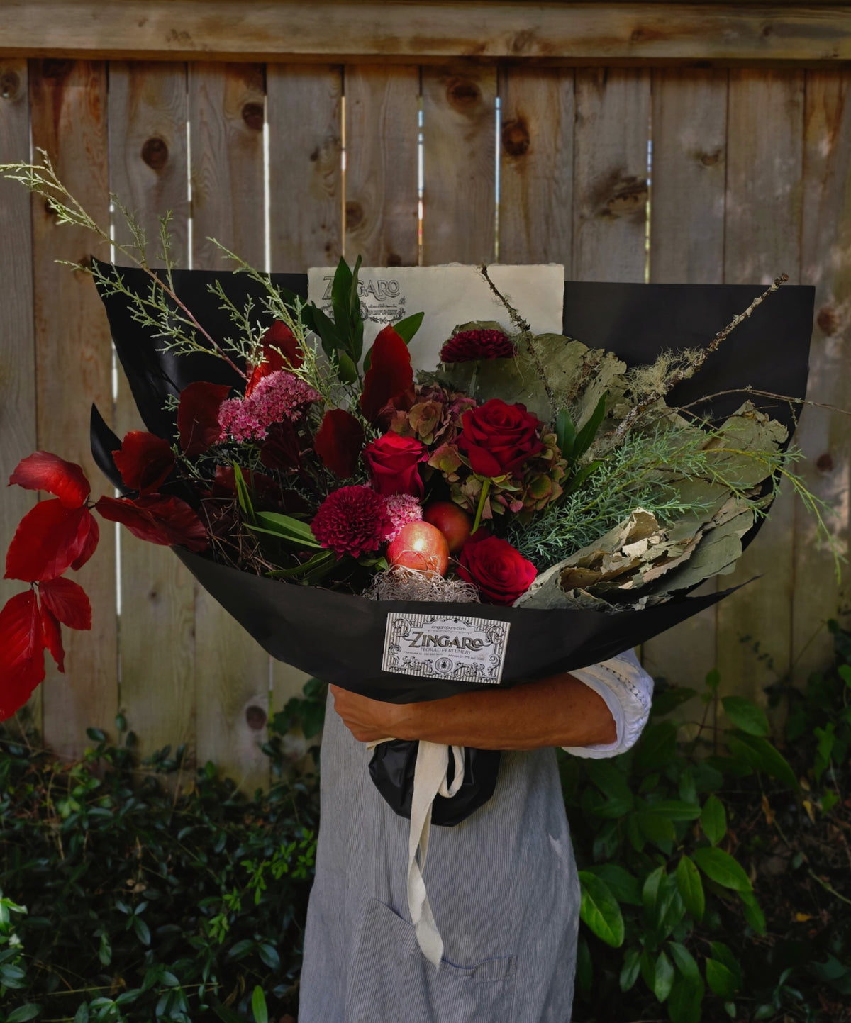 A large, beautifully arranged dried and fresh flower bouquet wrapped in black paper, held by a person wearing a gray apron in front of a wooden fence.