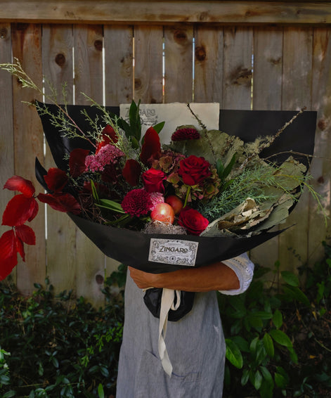A large, beautifully arranged dried and fresh flower bouquet wrapped in black paper, held by a person wearing a gray apron in front of a wooden fence.