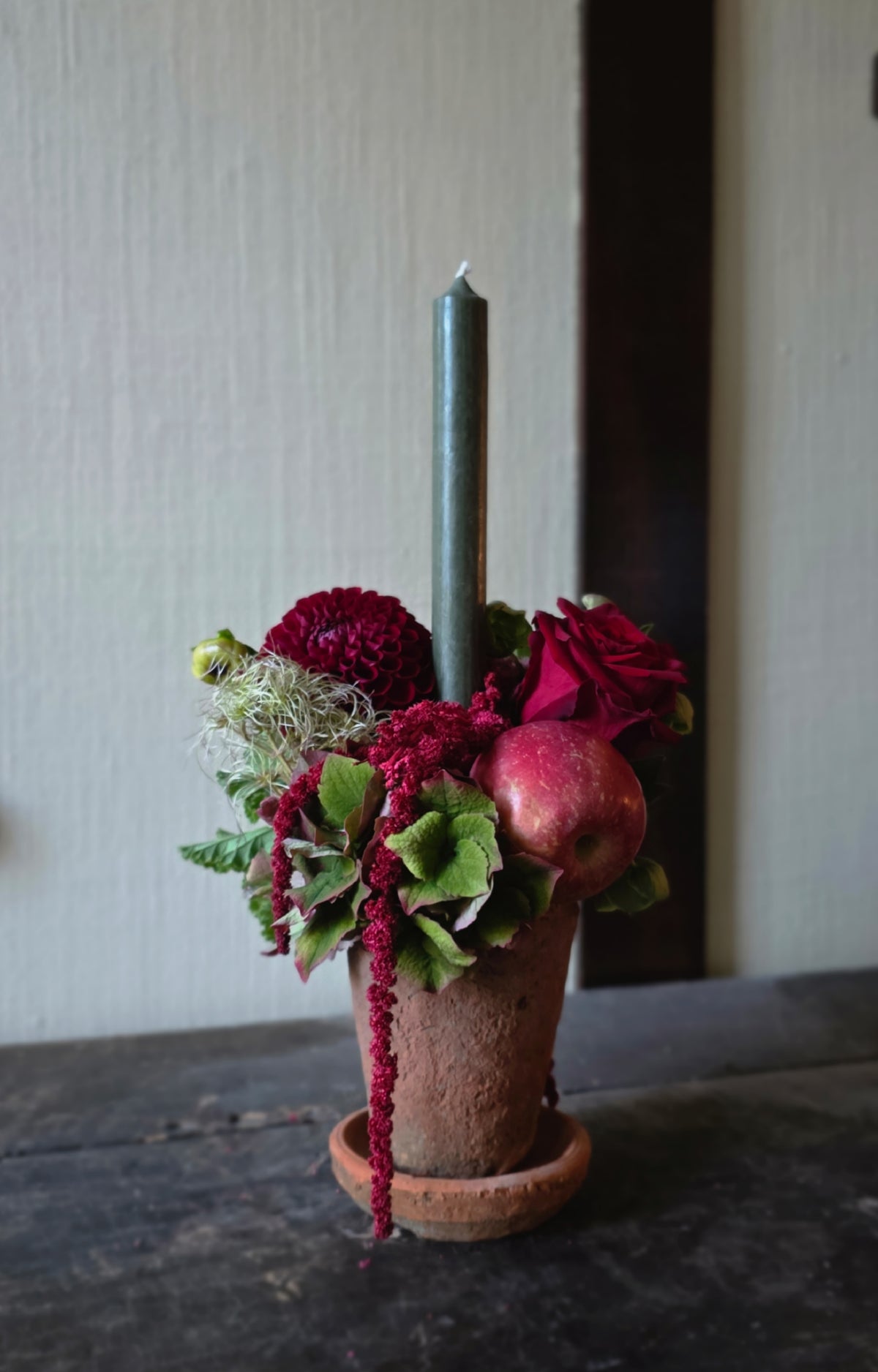 A rustic floral centerpiece in a terracotta pot with a tall dark green candle, deep red roses, burgundy amaryllis, green leaves, and a red apple on a dark wooden table against a light wall.