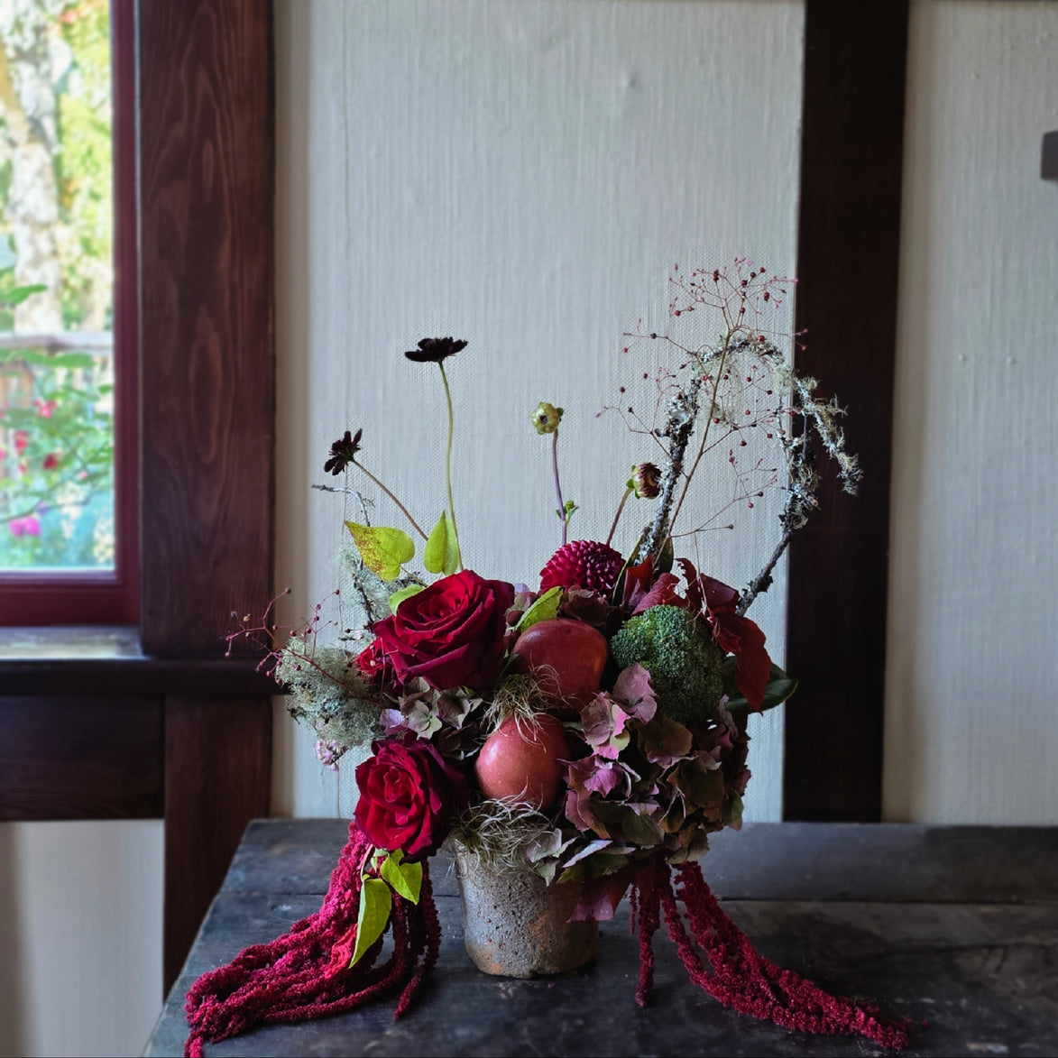 A rustic dried flower arrangement in a small textured pot, featuring deep red roses, burgundy berries, green foliage, and dried grasses, displayed on a dark wooden table in front of a white paneled wall and dark wood frame.