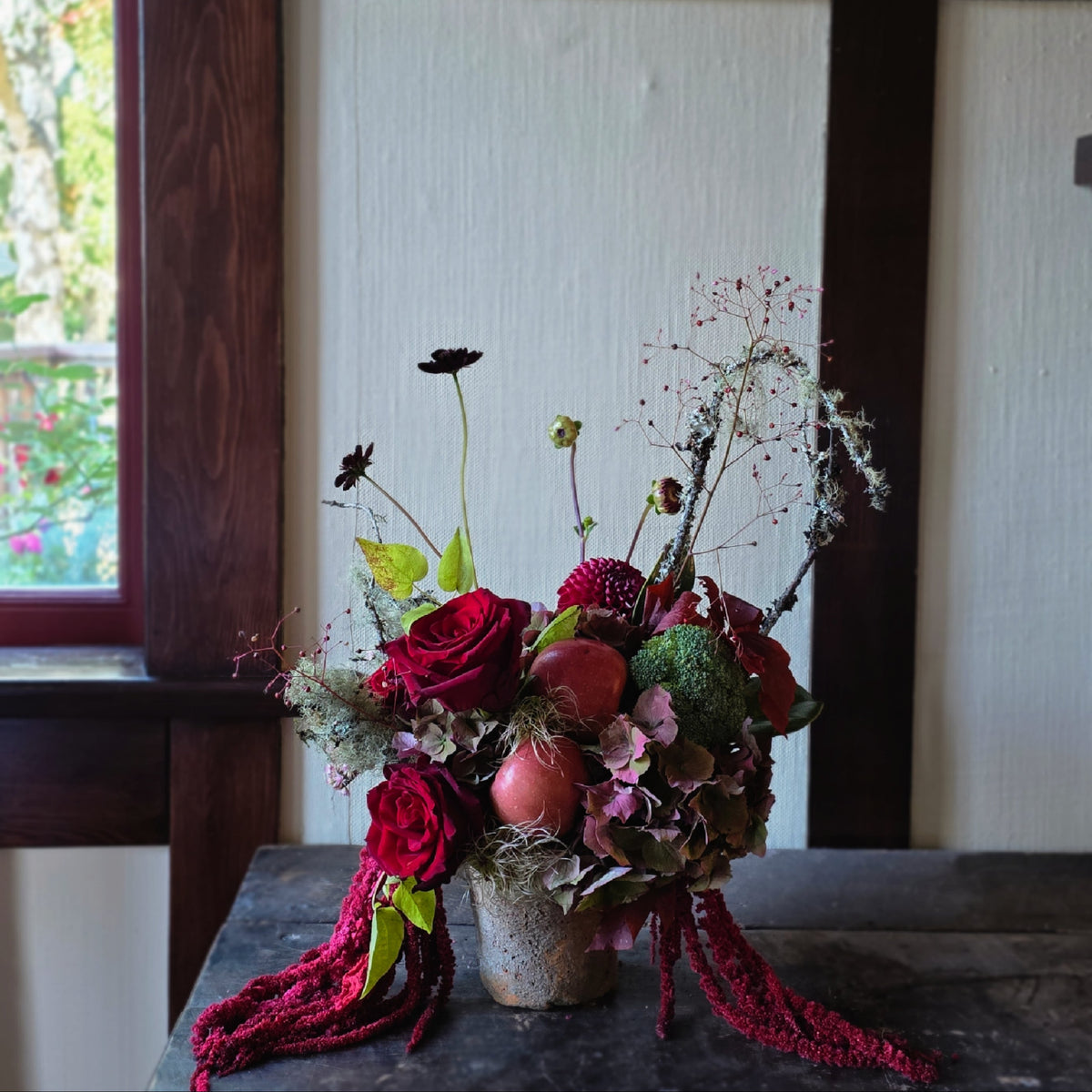 A rustic dried flower arrangement in a small textured pot, featuring deep red roses, burgundy berries, green foliage, and dried grasses, displayed on a dark wooden table in front of a white paneled wall and dark wood frame.