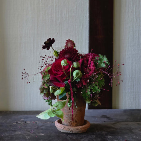 A rustic floral arrangement in a small terracotta pot with deep red roses, burgundy foliage, and green leaves, set on a dark wooden surface against a textured cream wall.