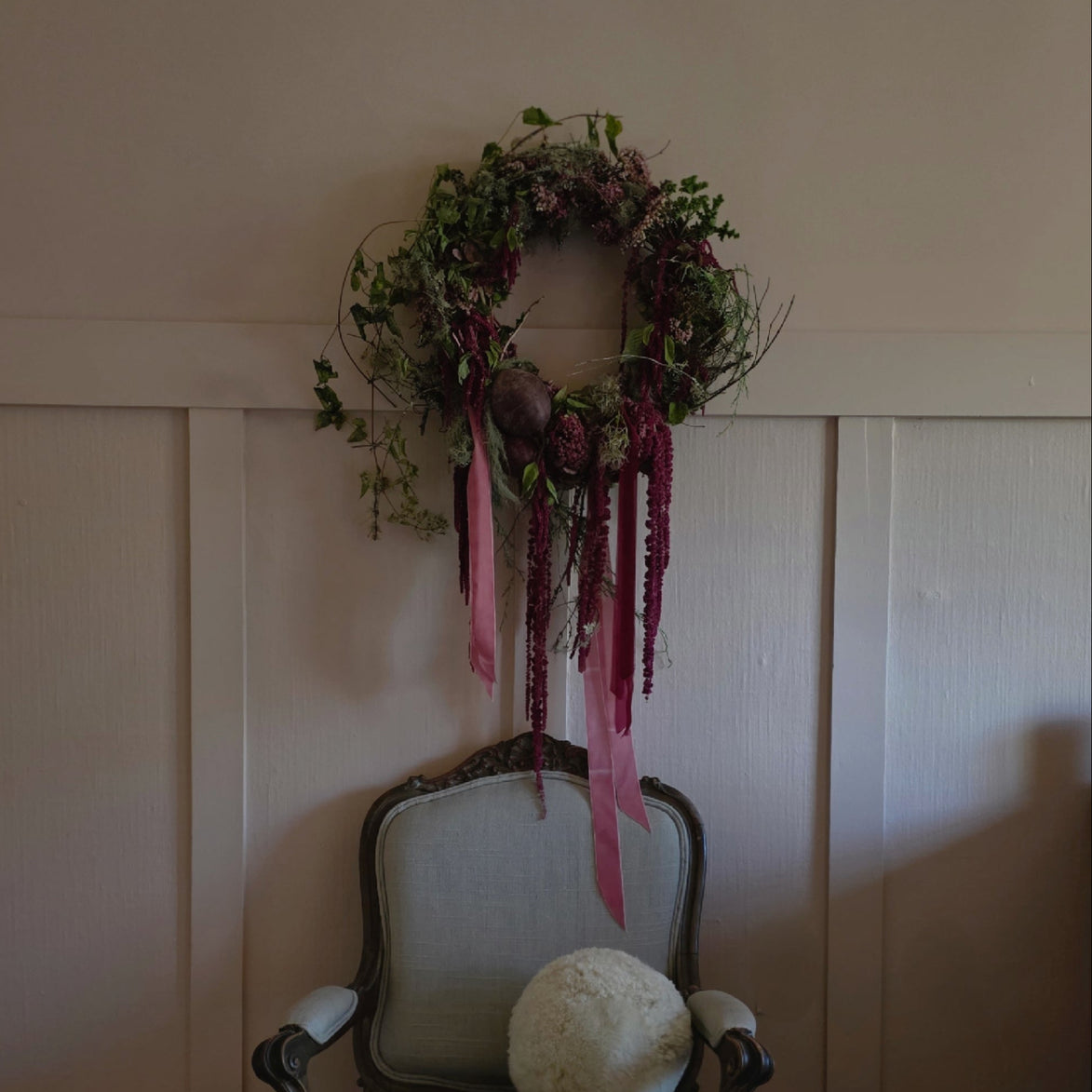 A decorative wreath with green foliage and pink hanging ribbons is displayed on a cream paneled wall above an antique-style upholstered armchair with a round cream pillow.