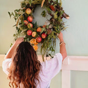 A woman with long wavy brown hair is seen from behind, hanging a large autumn-themed wreath on a pale green wall above a white railing. The wreath is decorated with green foliage, red apples, orange slices, and brown leaves.