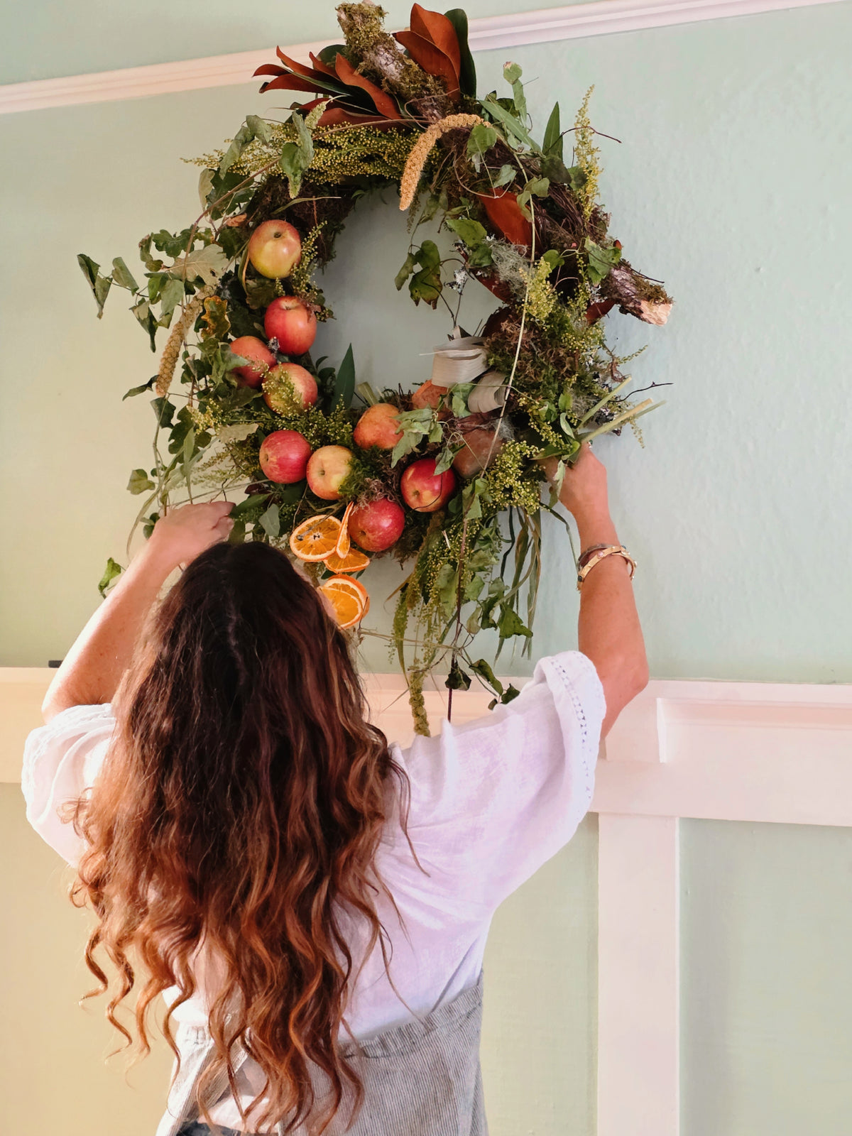 A woman with long wavy brown hair is seen from behind, hanging a large autumn-themed wreath on a pale green wall above a white railing. The wreath is decorated with green foliage, red apples, orange slices, and brown leaves.