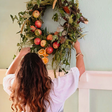 A woman with long wavy brown hair is seen from behind, hanging a large autumn-themed wreath on a pale green wall above a white railing. The wreath is decorated with green foliage, red apples, orange slices, and brown leaves.