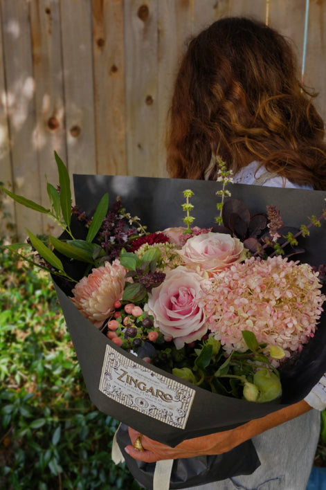 A person outdoors holding a large bouquet of pink and light pink flowers wrapped in black paper with a visible ZINGARO label. The background shows a wooden fence and greenery.