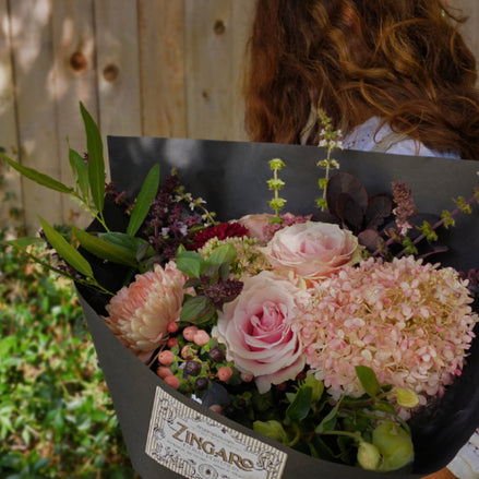 A person outdoors holding a large bouquet of pink and light pink flowers wrapped in black paper with a visible ZINGARO label. The background shows a wooden fence and greenery.